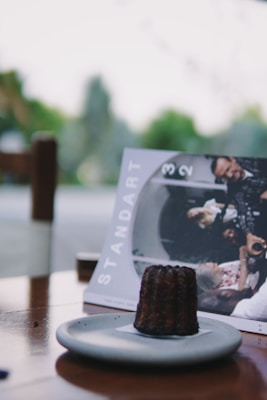 A close-up view of a dark, textured pastry on a light gray plate set on a wooden table. In the background, there is a magazine titled 'Standart' featuring a group of people engaged in a conversation. The setting appears to be outdoors with blurred greenery in the distance.