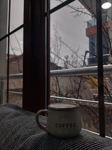 A cozy scene with a hand holding a warm ceramic coffee mug beside a window on a rainy day.