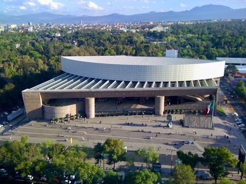 A large, modern building with a curved roof and a wide staircase area in front, set amidst a green, tree-filled urban environment. The architecture appears to be of a cultural or governmental facility. People can be seen walking around the plaza, which includes a prominent flag and a sculpture.