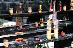 An assortment of candles in various glass containers, some decorated with Chinese characters, are placed on multi-tiered dark wooden shelves. Some candles appear melted, with wax dripped onto the shelves. The scene carries a traditional or religious ambiance, often seen in temples or places of worship.