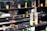 An assortment of candles in various glass containers, some decorated with Chinese characters, are placed on multi-tiered dark wooden shelves. Some candles appear melted, with wax dripped onto the shelves. The scene carries a traditional or religious ambiance, often seen in temples or places of worship.