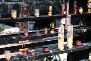 An assortment of candles in various glass containers, some decorated with Chinese characters, are placed on multi-tiered dark wooden shelves. Some candles appear melted, with wax dripped onto the shelves. The scene carries a traditional or religious ambiance, often seen in temples or places of worship.