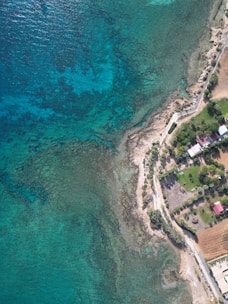 Aerial view of a coastal area featuring a dramatic shoreline with clear blue-green waters and rocky formations. The image shows a mix of residential buildings and greenery, including well-maintained lawns and gardens. There is a road running parallel to the shore, connecting the properties and surrounding areas. The coastal waters have varying shades of turquoise and deeper blue, indicating differing water depths.
