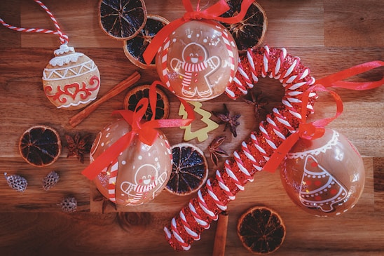 Festive ornaments and decorations are arranged on a wooden surface. Gingerbread-themed baubles with red ribbons, a large candy cane style decoration, and dried citrus slices create a holiday atmosphere. Cinnamon sticks and star anise add warmth and spice to the scene.