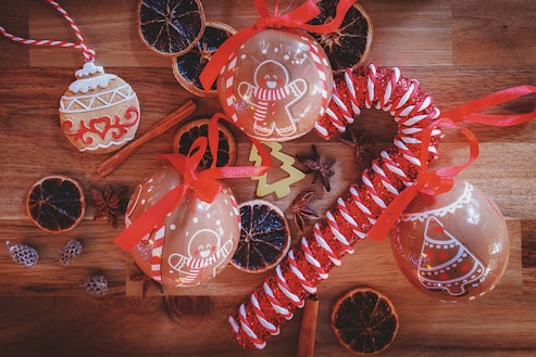 Festive ornaments and decorations are arranged on a wooden surface. Gingerbread-themed baubles with red ribbons, a large candy cane style decoration, and dried citrus slices create a holiday atmosphere. Cinnamon sticks and star anise add warmth and spice to the scene.