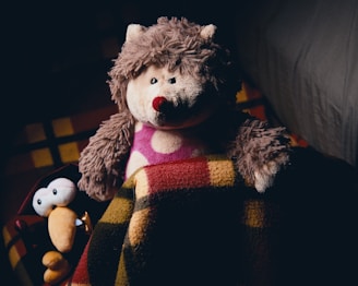 A plush hedgehog toy with a red nose and pink polka dot fabric on its body is sitting next to a smaller cartoonish stuffed bird with large, cartoonish eyes and an orange beak. Both are tucked under a multi-colored checkered blanket, on a bed.