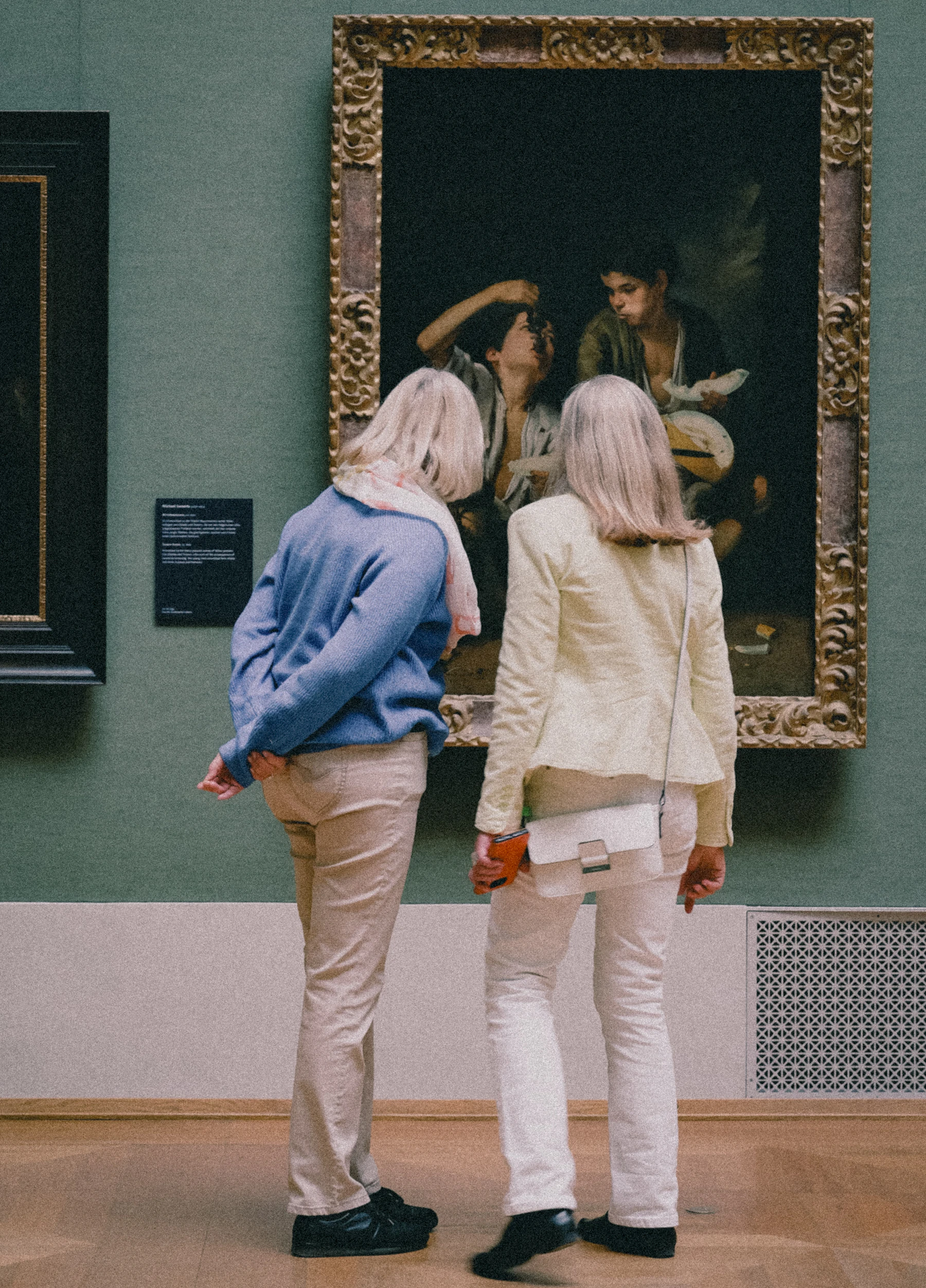 Two visitors seen from behind standing close to a dark painting in an ornate gilt frame on a sage green gallery wall