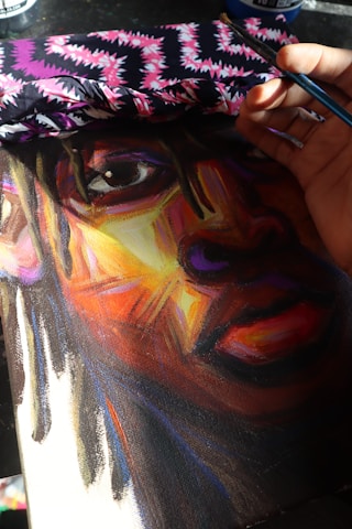 A close-up view of a vibrant, colorful painting of a person's face, partially covered with a piece of fabric displaying a bold, zigzag pattern in black, white, and pink. The artist's hand, holding a paintbrush, is seen applying finishing touches to the artwork.