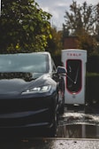 A black electric car is parked at a charging station with visible greenery and wet ground reflections. The charging unit is prominently branded behind the vehicle.
