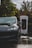 A black electric car is parked at a charging station with visible greenery and wet ground reflections. The charging unit is prominently branded behind the vehicle.