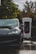 A black electric car is parked at a charging station with visible greenery and wet ground reflections. The charging unit is prominently branded behind the vehicle.