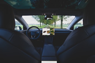 The image captures the interior of a modern car, looking out from the backseat towards the windshield. A central display screen shows navigation or controls, and the surroundings outside include a green, tree-lined road.