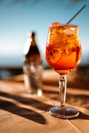 A glass of Aperol Spritz filled with ice cubes is placed prominently on a table against a blurred background. The sunlight creates a warm glow, enhancing the vibrant orange hue of the drink. A bottle is slightly visible in the background, adding to the aesthetic of a refreshing outdoor setting.