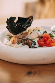 A gourmet dish featuring a combination of seafood and fresh ingredients. Plump shrimp, grilled calamari, and octopus are arranged with a scoop of creamy sauce, topped with green herbs. The dish is garnished with a black crispy element and accompanied by diced red tomatoes, creating a visually striking presentation.