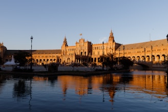 An expansive architectural structure reflecting a combination of Renaissance and Moorish styles, prominently illuminated by the warm glow of a setting sun. A tranquil body of water reflects the building's intricate details and the clear sky above. Lamp posts and trees dot the well-maintained surroundings, adding to the scene's charm and historical ambiance.