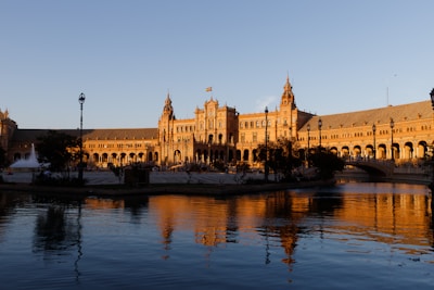 An expansive architectural structure reflecting a combination of Renaissance and Moorish styles, prominently illuminated by the warm glow of a setting sun. A tranquil body of water reflects the building's intricate details and the clear sky above. Lamp posts and trees dot the well-maintained surroundings, adding to the scene's charm and historical ambiance.