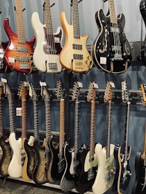 A collection of electric and bass guitars hanging on display racks. The guitars vary in color, shape, and design, showcasing a range of wood finishes and hardware styles.