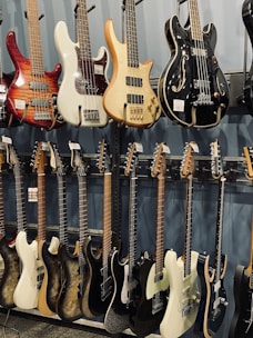 A collection of electric and bass guitars hanging on display racks. The guitars vary in color, shape, and design, showcasing a range of wood finishes and hardware styles.