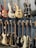 A collection of electric and bass guitars hanging on display racks. The guitars vary in color, shape, and design, showcasing a range of wood finishes and hardware styles.