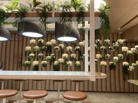 A modern interior space features a wall adorned with vertical cylindrical elements, each topped with lush greenery. Above, large pendant lights hang over a row of bar stools with brown cushioned seats. The environment suggests a blend of natural and contemporary design elements.