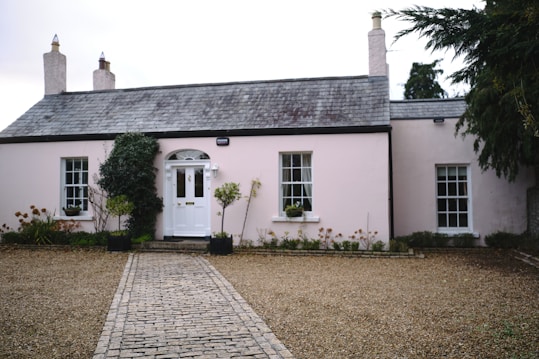 A quaint cream-colored cottage with a slate roof is surrounded by a neatly organized garden and a gravel driveway. The building features chimney stacks, large sash windows with white frames, and a central white door with decorative arch window above. Potted plants and shrubs add greenery to the setting.