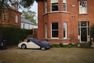 A vintage car is partially covered with a tarp next to a brick house with large windows. The house features a dark-colored front door with the number 72. There are bushes and plants bordering the pathway in the foreground. Another brick building and some greenery are visible in the background.
