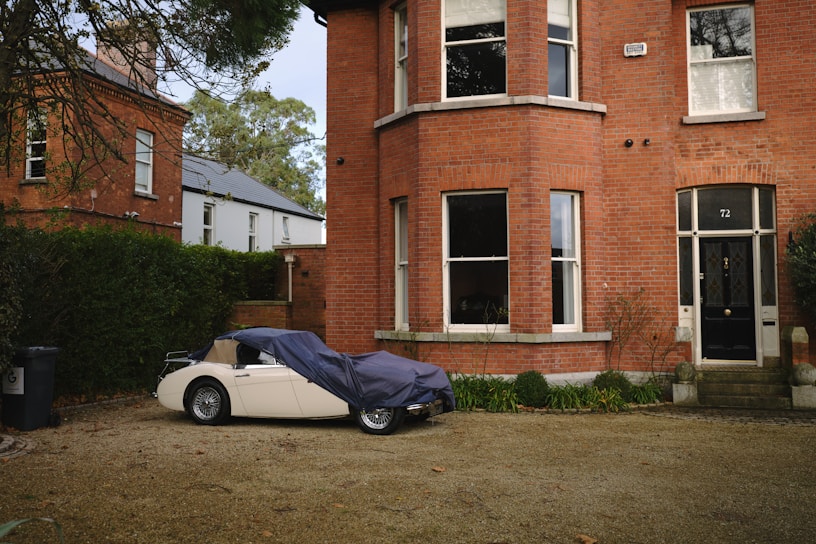A vintage car is partially covered with a tarp next to a brick house with large windows. The house features a dark-colored front door with the number 72. There are bushes and plants bordering the pathway in the foreground. Another brick building and some greenery are visible in the background.