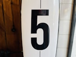 A large black numeral '5' is prominently displayed on a white background. To the left, there's a wooden surface with visible grain patterns, and to the right, a section of white tiled wall is partially visible.