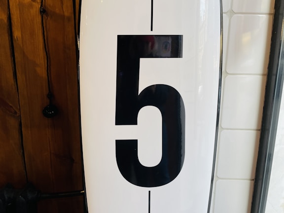 A large black numeral '5' is prominently displayed on a white background. To the left, there's a wooden surface with visible grain patterns, and to the right, a section of white tiled wall is partially visible.