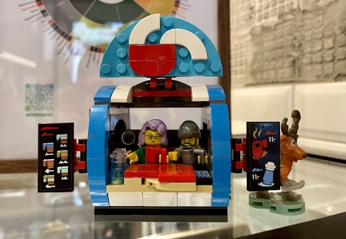 A LEGO toy set depicting what appears to be a small cafe or food stall with two minifigures inside. One minifigure has lavender hair and glasses, while the other wears a helmet. The front of the stall features open doors with graphics of drinks, and a small reindeer figurine is placed next to it. The stall is built with colorful bricks including blue, white, red, and blue-grey tones.