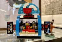 A LEGO toy set depicting what appears to be a small cafe or food stall with two minifigures inside. One minifigure has lavender hair and glasses, while the other wears a helmet. The front of the stall features open doors with graphics of drinks, and a small reindeer figurine is placed next to it. The stall is built with colorful bricks including blue, white, red, and blue-grey tones.