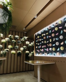 A modern interior space featuring a wall with cylindrical planters filled with lush green plants. The adjacent wall displays a geometric pattern of colorful circular disks illuminated by backlighting, creating a striking visual effect. The setting includes minimalistic tables with light-colored surfaces, enhancing the contemporary design.