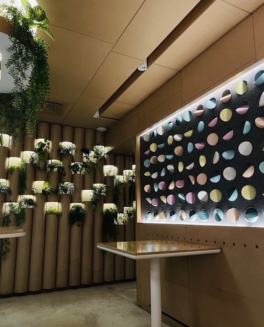 A modern interior space featuring a wall with cylindrical planters filled with lush green plants. The adjacent wall displays a geometric pattern of colorful circular disks illuminated by backlighting, creating a striking visual effect. The setting includes minimalistic tables with light-colored surfaces, enhancing the contemporary design.