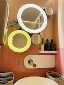 A modern bathroom setup features a stylish wall with circular mirrors of varying sizes, two of which have integrated lights glowing in white and yellow. Below the mirrors, there is a shelf holding two green bottles and a green toothbrush. A sleek faucet extends over a white basin, with folded towels and baskets neatly arranged nearby.