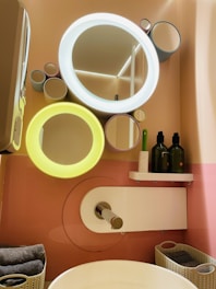 A modern bathroom setup features a stylish wall with circular mirrors of varying sizes, two of which have integrated lights glowing in white and yellow. Below the mirrors, there is a shelf holding two green bottles and a green toothbrush. A sleek faucet extends over a white basin, with folded towels and baskets neatly arranged nearby.