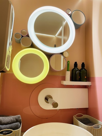 A modern bathroom setup features a stylish wall with circular mirrors of varying sizes, two of which have integrated lights glowing in white and yellow. Below the mirrors, there is a shelf holding two green bottles and a green toothbrush. A sleek faucet extends over a white basin, with folded towels and baskets neatly arranged nearby.