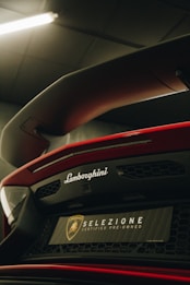 A close-up of the rear end of a red Lamborghini featuring a prominent rear wing and branding. The image is taken in a dimly lit environment, highlighting the aerodynamic features and sleek design. The 'Selezione Certified Pre-Owned' badge is visible, suggesting a focus on exclusivity and luxury.