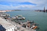A waterfront scene featuring a bustling dock with multiple boats, a serene expanse of water stretching into the distance, and a cityscape on the horizon. The dock is lined with small boats and water taxis. Numerous people are walking along the pier. To the right, a tower structure stands out, possibly part of a historical building.