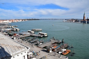 A waterfront scene featuring a bustling dock with multiple boats, a serene expanse of water stretching into the distance, and a cityscape on the horizon. The dock is lined with small boats and water taxis. Numerous people are walking along the pier. To the right, a tower structure stands out, possibly part of a historical building.