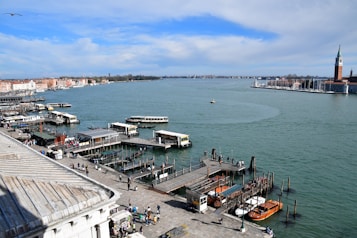 A waterfront scene featuring a bustling dock with multiple boats, a serene expanse of water stretching into the distance, and a cityscape on the horizon. The dock is lined with small boats and water taxis. Numerous people are walking along the pier. To the right, a tower structure stands out, possibly part of a historical building.