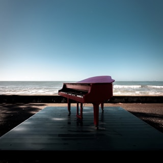 A red piano is placed on a platform by the sea. The ocean waves are gently rolling in the background, and the clear sky creates a serene atmosphere.