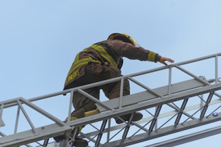 A firefighter dressed in a protective suit and helmet climbs a metal ladder extending upwards. The background features a clear blue sky.