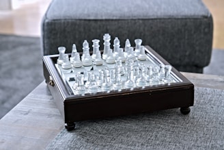 A glass chess set is arranged on a square wooden board with ornate edging, placed on a textured grey fabric surface. The chess pieces are clear and white, reflecting light in a subtly elegant manner.