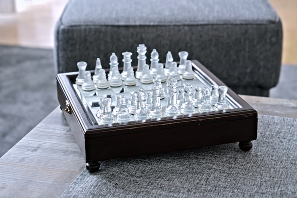 A glass chess set is arranged on a square wooden board with ornate edging, placed on a textured grey fabric surface. The chess pieces are clear and white, reflecting light in a subtly elegant manner.