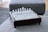 A glass chess set is arranged on a square wooden board with ornate edging, placed on a textured grey fabric surface. The chess pieces are clear and white, reflecting light in a subtly elegant manner.