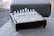 A glass chess set is arranged on a square wooden board with ornate edging, placed on a textured grey fabric surface. The chess pieces are clear and white, reflecting light in a subtly elegant manner.