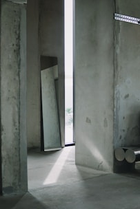 A minimalist interior space featuring concrete walls and floors. A narrow strip of light enters through a tall, narrow window, casting shadows on the floor. A large mirror rests against the wall, reflecting part of the room. Metal beams or pipes are positioned in the corner, adding an industrial touch to the setting.