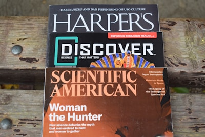 Three science magazines are stacked on a wooden surface. The top magazine is Harper's, featuring articles on UFO culture. Below it is Discover, with a focus on exposing research fraud and other scientific topics, including space. The bottom magazine, Scientific American, has a cover story titled 'Woman the Hunter,' challenging traditional gender roles in evolution. It also includes topics on organ transplants and endangered species.