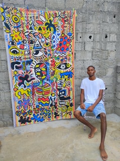 A man sits next to a vibrant, colorful abstract painting mounted on a concrete wall. The artwork features numerous geometric shapes, abstract faces, and eye motifs in a mix of bold colors including red, blue, yellow, and black, creating a visually stimulating pattern.