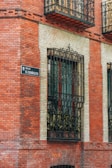 A brick building facade with ornate black wrought iron window grills. A decorative street sign reads 'Calle de El Españoleto.' The windows are bordered by light-colored bricks, contrasting with the red brick exterior.